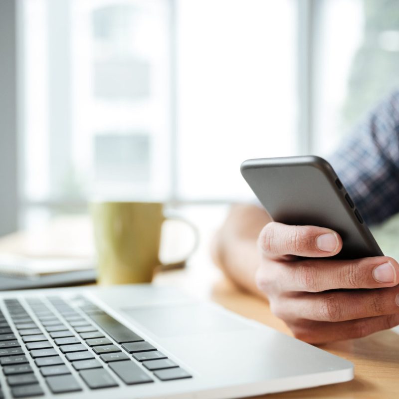 Cropped image of young man sitting in office coworking while using laptop computer and mobile phone.